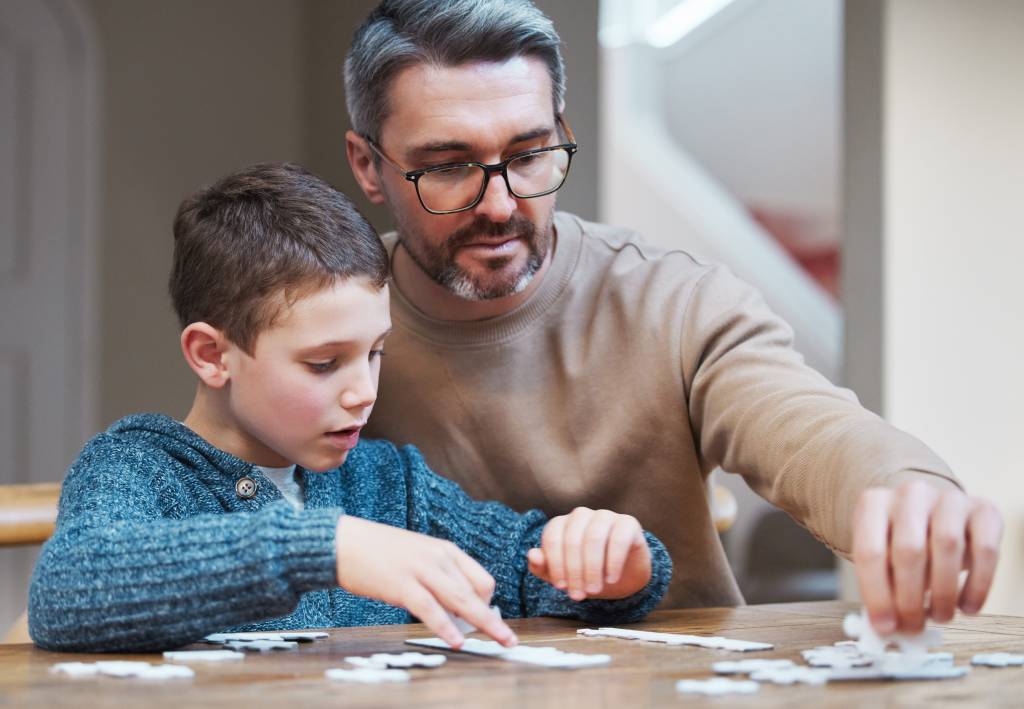 Adult and child assembling jigsaw puzzle together at wooden table during supportive interaction